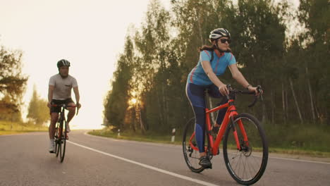 Two-cyclists-a-man-and-a-woman-ride-on-the-highway-on-road-bikes-wearing-helmets-and-sportswear-at-sunset-in-slow-motion